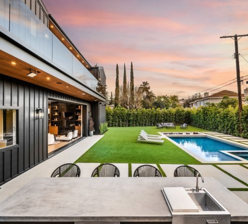 Modern backyard with a swimming pool, outdoor kitchen, and lounge area at sunset.